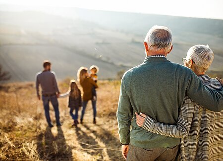 HWF Titelbild Senioren Ein älteres Paar, Mann und Frau, haben jeweils den Arm um den anderen gelegt, sind nur von hinten fotografiert und sehen, auf einem Trockenhang stehend ins Tal. Vor Ihnen befindet sich eine Familie, Mann, Frau und zwei Kinder.